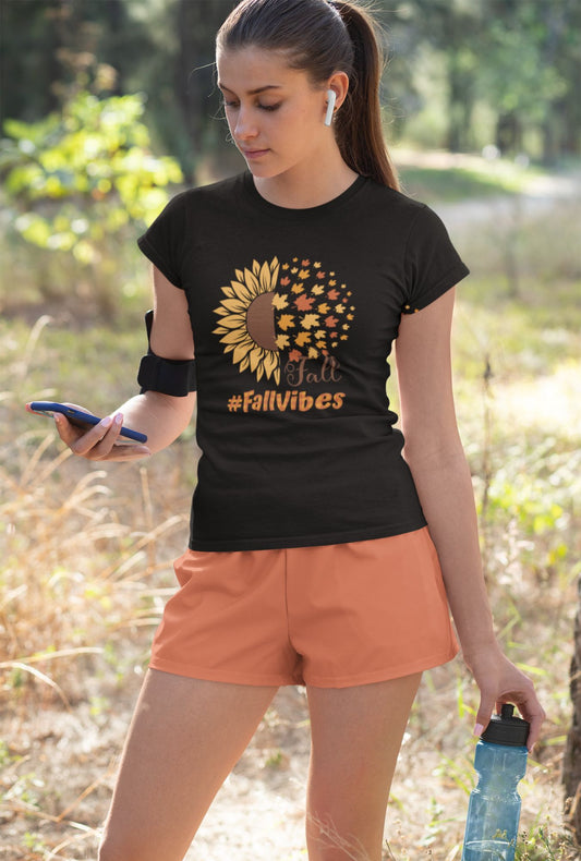 A woman wearing a RedBerry Cuddle 'Fall Vibes' Women's T-Shirt, made of 100% cotton. The black shirt features a sunflower graphic transitioning into autumn leaves with the words 'Fall' and '#FallVibes' in warm, autumnal colors.