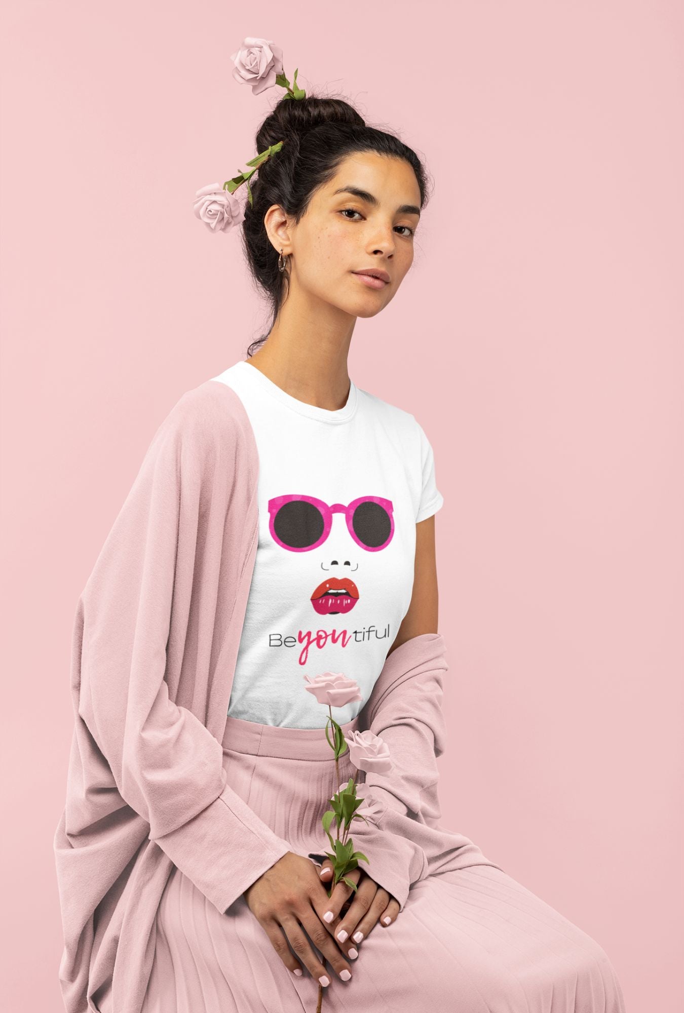 A woman wearing a RedBerry Cuddle 'Beautiful' Women's T-Shirt made of 100% cotton. The shirt features a design with pink sunglasses, red lips, and the word 'BeYOUtiful' printed on it. She is styled with pink roses in her hair and is draped in a pink shawl, holding a pink rose against a matching pink background.