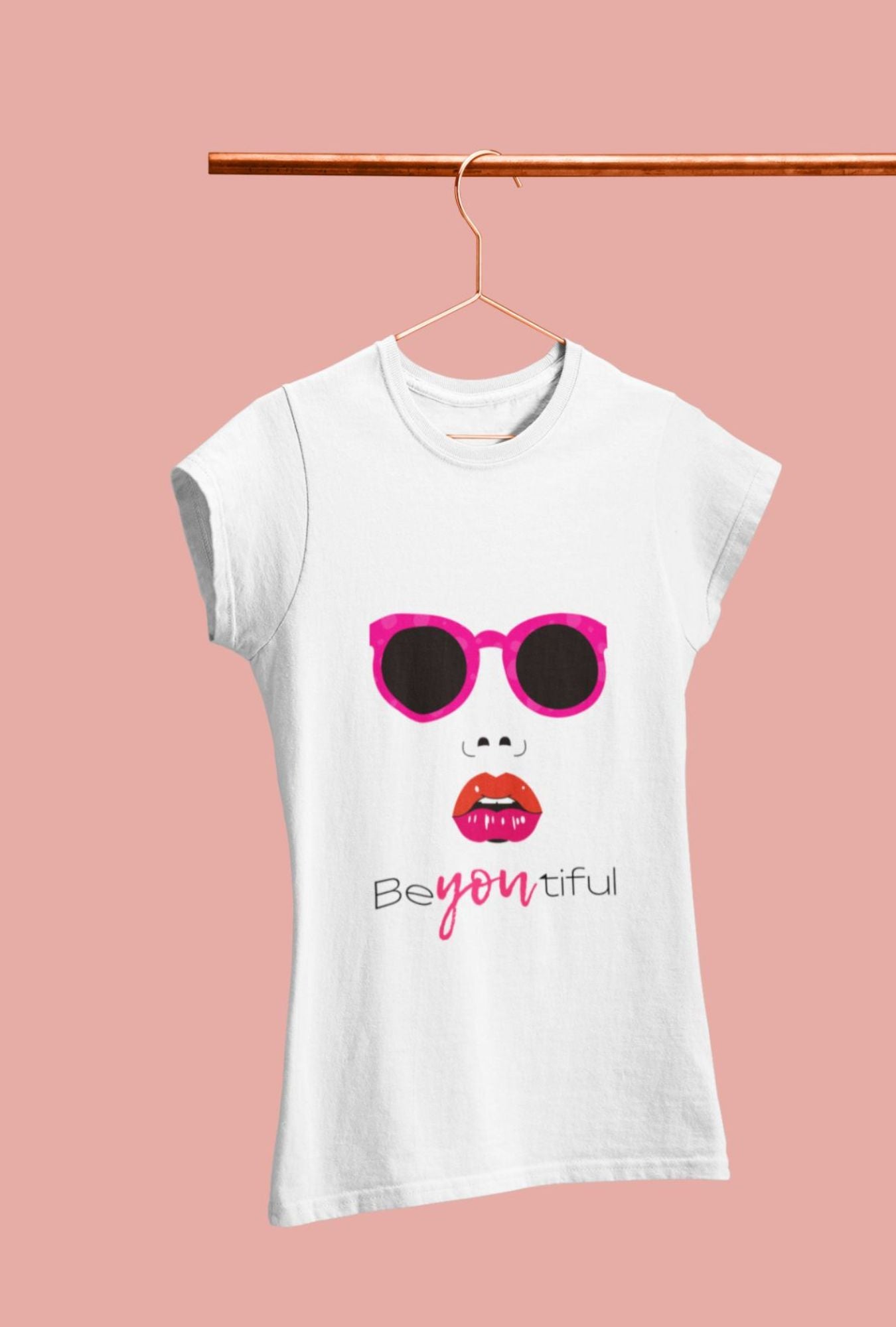 A woman wearing a RedBerry Cuddle 'Beautiful' Women's T-Shirt made of 100% cotton. The shirt features a design with pink sunglasses, red lips, and the word 'BeYOUtiful' printed on it. She is styled with pink roses in her hair and is draped in a pink shawl, holding a pink rose against a matching pink background.